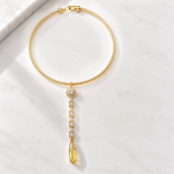 A gold Y-necklace featuring a textured chain, a central round bezel-set clear stone, and a dangling teardrop-shaped yellow gemstone, displayed on a white marble surface.