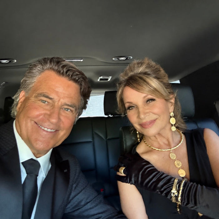 A man in a dark suit and tie and a woman in a black dress with long black gloves and gold jewelry sitting together inside a vehicle.