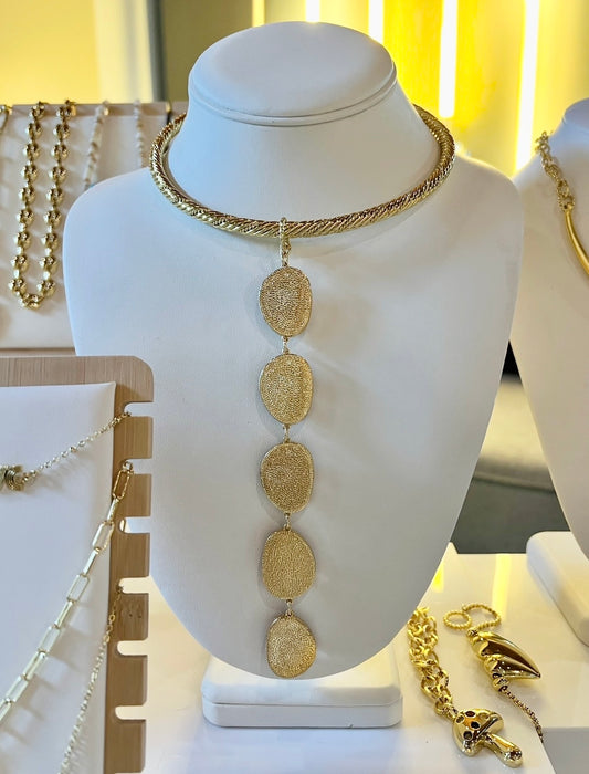 A gold-toned necklace displayed on a white jewelry bust. The necklace features a thick braided choker band and five vertically aligned oval links with textured, hammered finishes.