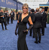 A woman wearing a floor-length black gown with a plunging neckline, long gloves, and a gold necklace stands on a blue carpet at an Emmy event, with photographers and guests in the background.