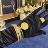 A black satin glove displays a gold Emmy statuette ring and matching gold cuff bracelets on a blue carpet with an Emmys backdrop.