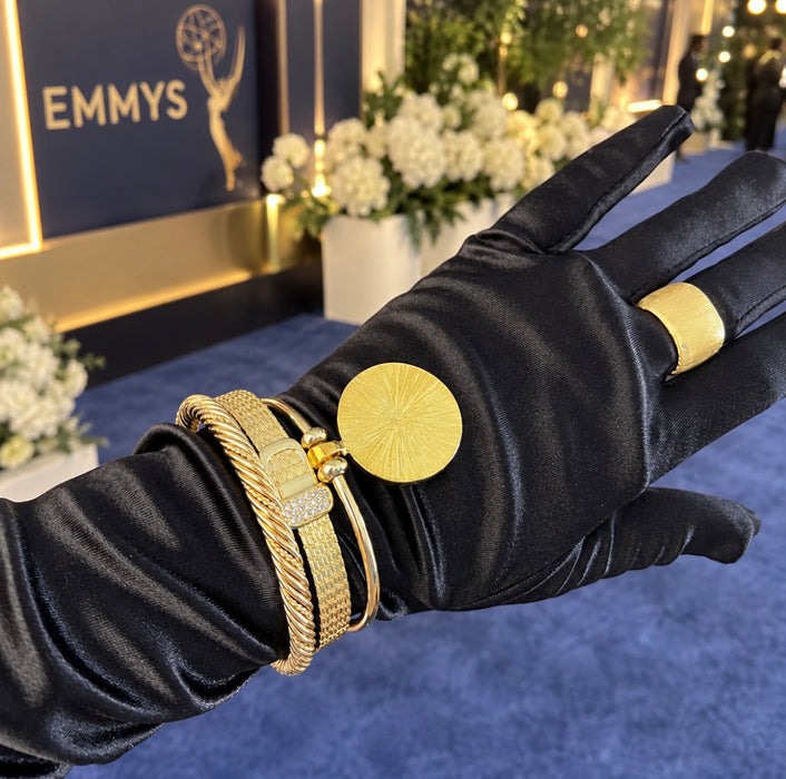 A black satin glove displays a gold Emmy statuette ring and matching gold cuff bracelets on a blue carpet with an Emmys backdrop.