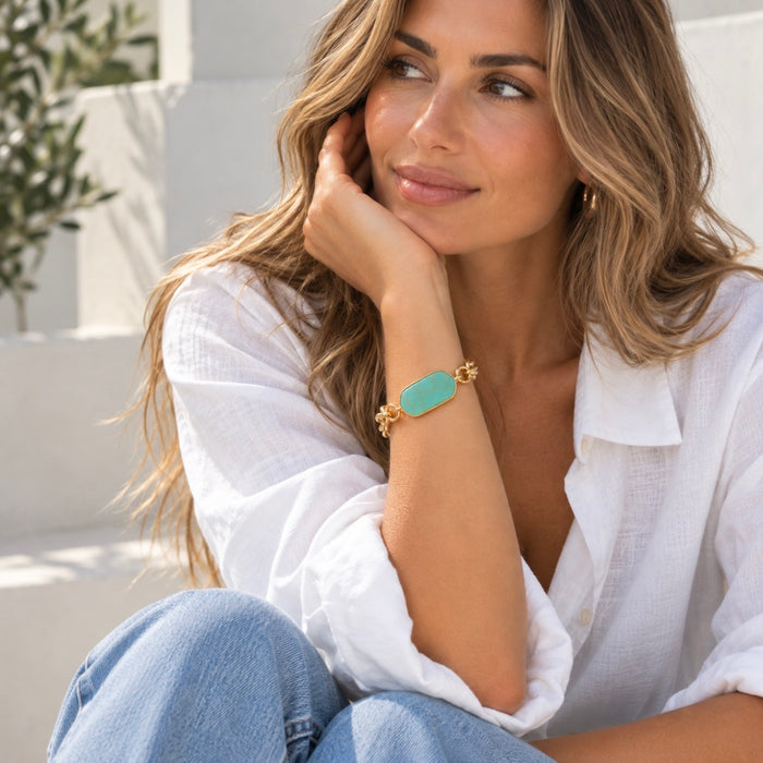 A woman wearing a gold bracelet with a mint green stone, posing in a white shirt and blue jeans.