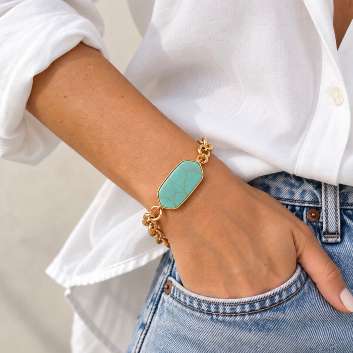 A close-up of a person's wrist wearing a gold chain bracelet with a turquoise-colored stone centerpiece, styled with a white button-down shirt and blue jeans.
