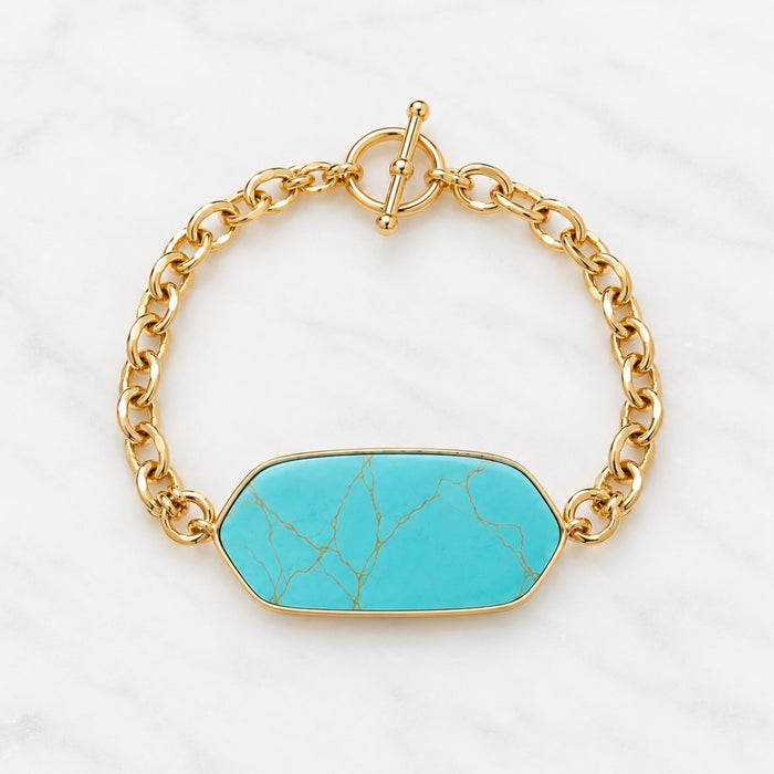 A gold chain bracelet with a hexagonal turquoise stone centerpiece, displayed on a white marble background.
