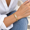 A close-up of a person's wrist wearing a gold mesh bracelet with a rectangular gold clasp and a clear crystal-encrusted center, styled with a white button-down shirt and blue jeans.