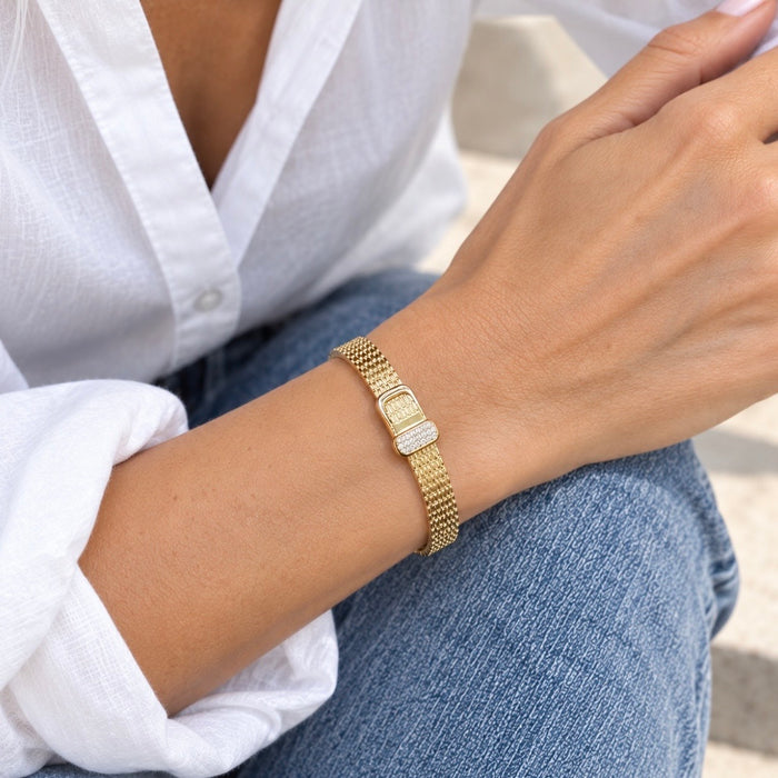 A close-up of a person's wrist wearing a gold mesh bracelet with a rectangular gold clasp and a clear crystal-encrusted center, styled with a white button-down shirt and blue jeans.