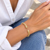 A close-up of a person's wrist wearing a delicate gold cable-style bracelet with two small ball accents, paired with a white button-up shirt and blue jeans.