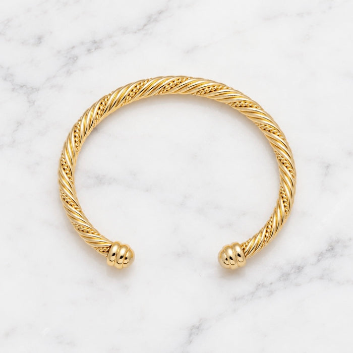 A gold cable-wrapped cuff bracelet with polished ball-end closures, displayed on a white marble surface.