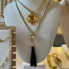 A gold-tone layered necklace displayed on a white mannequin bust, featuring a black tassel pendant with a clear rhinestone and a circular gold medallion, with other jewelry visible in the background.