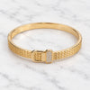 A gold bangle bracelet with a textured mesh band and a rectangular buckle embellished with small clear stones, displayed on a white marble surface.