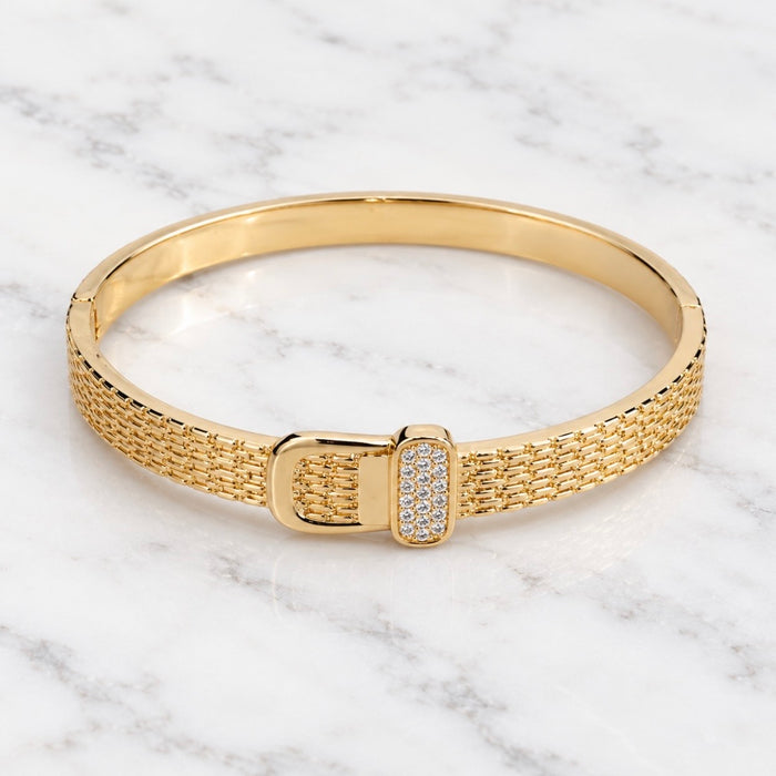 A gold bangle bracelet with a textured mesh band and a rectangular buckle embellished with small clear stones, displayed on a white marble surface.