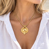 A gold pendant necklace with a circular sunburst design worn by a model in a white blouse.