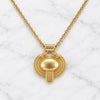 A gold pendant necklace featuring a stylized, textured circular medallion with a central dome and a vertical bar, suspended from a delicate gold chain on a white marble background.