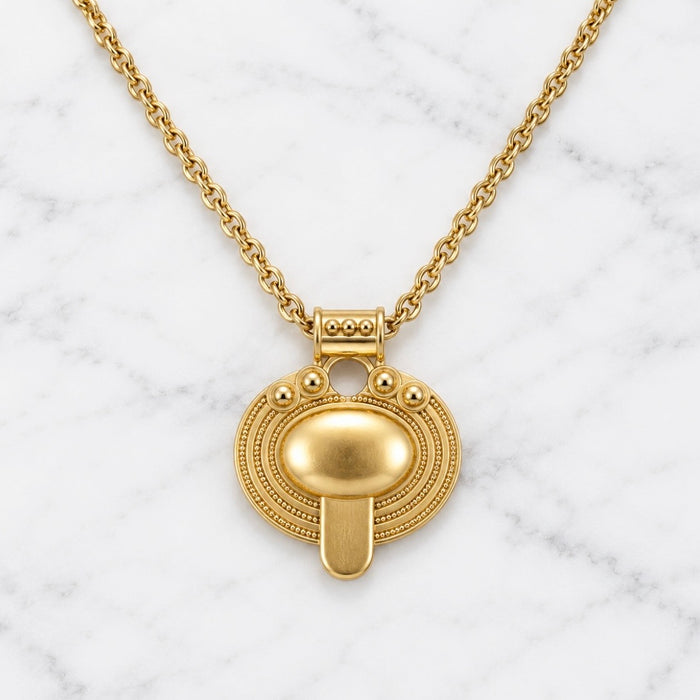 A gold pendant necklace featuring a stylized, textured circular medallion with a central dome and a vertical bar, suspended from a delicate gold chain on a white marble background.
