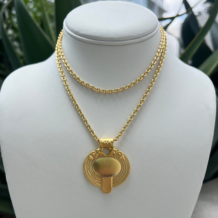 A gold double-strand necklace with a mushroom-shaped pendant, displayed on a white jewelry bust with blurred green foliage in the background.
