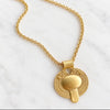 A gold pendant necklace featuring a circular textured medallion with a central heart-shaped pendant, displayed on a white marble surface.