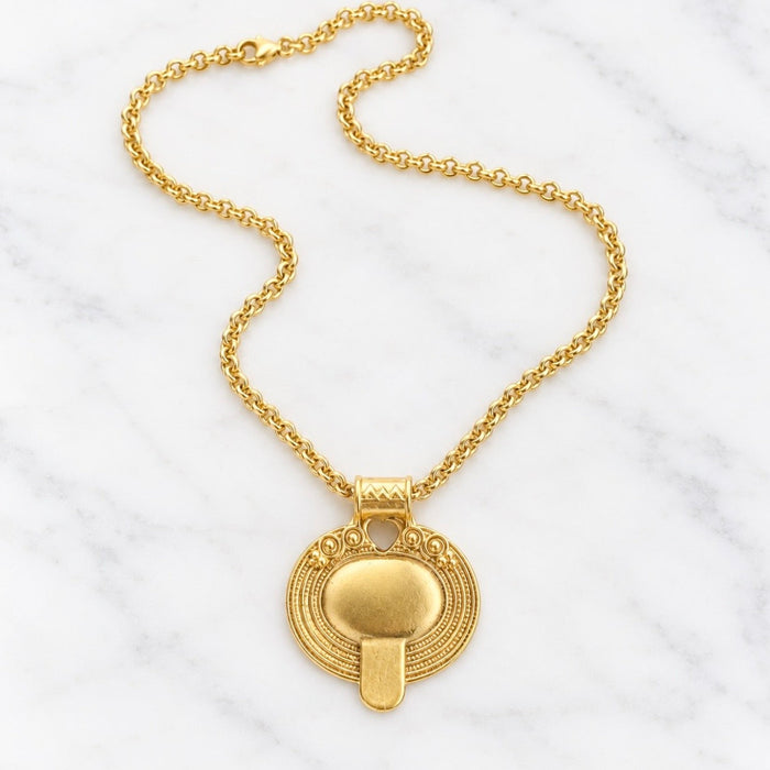 A gold pendant necklace featuring a textured circular medallion with a central vertical bar, displayed on a white marble background.