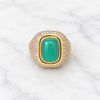 A gold signet ring featuring a large, smooth green cabochon gemstone in the center, surrounded by a halo of small pavé-set clear stones on a white marble background.