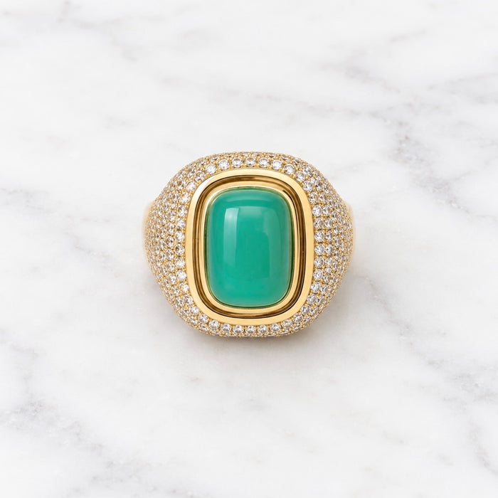 A gold signet ring featuring a large, smooth green cabochon gemstone in the center, surrounded by a halo of small pavé-set clear stones on a white marble background.
