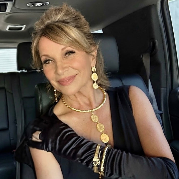 A woman seated inside a black car wearing a black sleeveless top, long black satin gloves, layered gold coin necklaces, matching drop earrings, and gold bracelets.