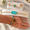 A lifestyle shot of a gold bracelet with a turquoise stone worn on a woman's wrist, displayed alongside other jewelry pieces on white display stands over a marble surface.