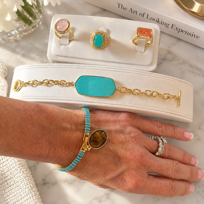 A lifestyle shot of a gold bracelet with a turquoise stone worn on a woman's wrist, displayed alongside other jewelry pieces on white display stands over a marble surface.