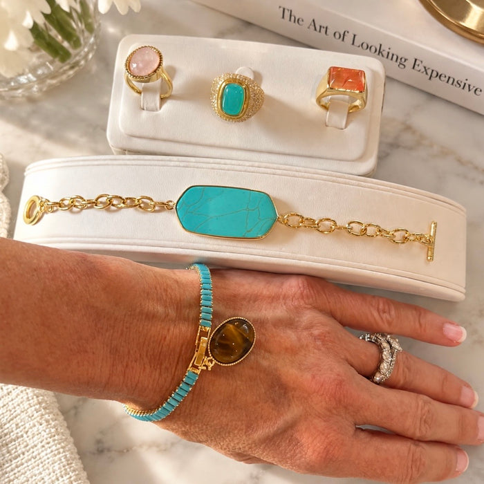 A hand wearing a turquoise and gold beaded bracelet with a large oval stone, displayed next to a matching bracelet laid on a cream jewelry display. The scene also shows gold rings on white display cards, with a marble surface and a book in the background.