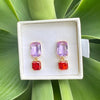 A pair of gold-tone drop earrings with a light purple rectangular stone on top and a textured red square stone below, displayed in a white jewelry box on a bed of green leaves.