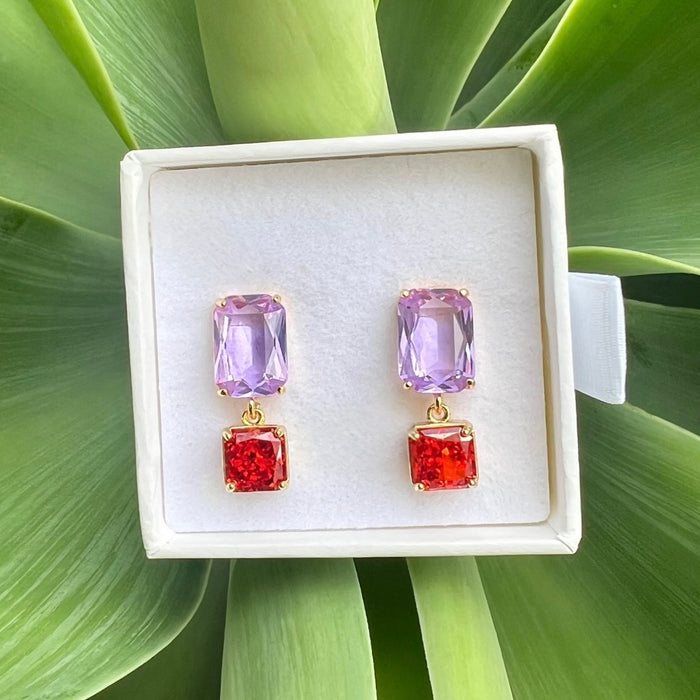 A pair of gold-tone drop earrings with a light purple rectangular stone on top and a textured red square stone below, displayed in a white jewelry box on a bed of green leaves.