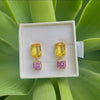 A pair of gold drop earrings with a yellow top stone and a pink glitter bottom stone, presented in a white jewelry box on a background of green leaves.