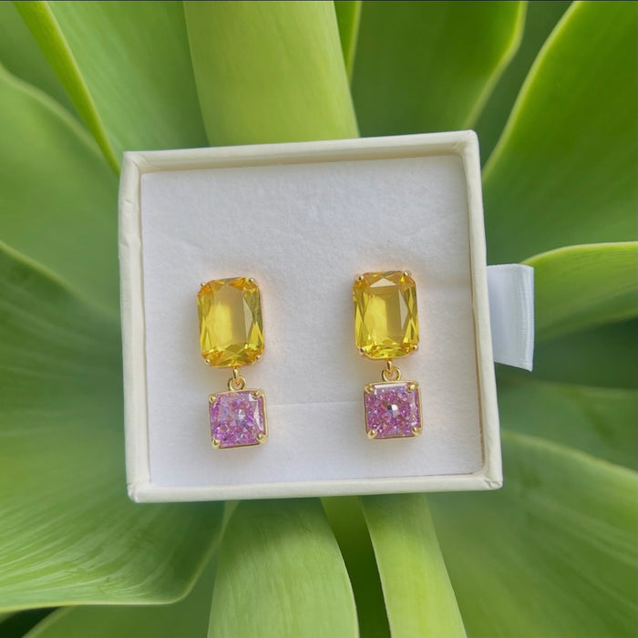 A pair of gold drop earrings with a yellow top stone and a pink glitter bottom stone, presented in a white jewelry box on a background of green leaves.