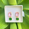 A pair of gold-tone drop earrings with rectangular pink stones on top and square green stones below, displayed in a white jewelry box against a bright green leaf background.