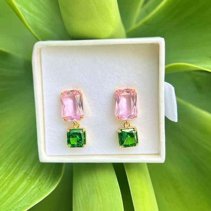 A pair of gold-tone drop earrings with rectangular pink stones on top and square green stones below, displayed in a white jewelry box against a bright green leaf background.