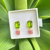 A pair of gold drop earrings with green rectangular top stones and pink square bottom stones, displayed in a white jewelry box on a green leaf background.