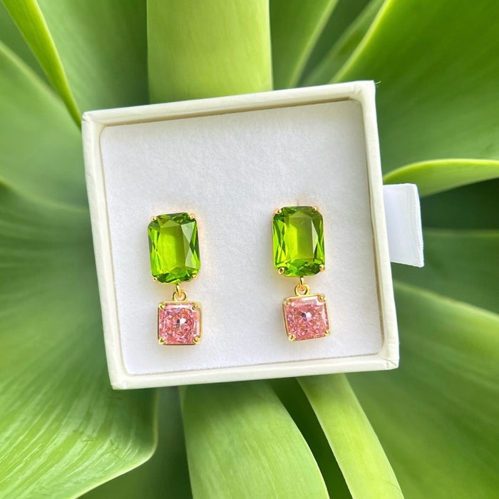 A pair of gold drop earrings with green rectangular top stones and pink square bottom stones, displayed in a white jewelry box on a green leaf background.