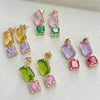 Four pairs of gold-tone double-drop dangle earrings with rectangular and square-cut gemstones in yellow, green, purple, and red, arranged on a light gray textured background.