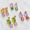 Four pairs of gold-tone drop earrings with rectangular gemstones in yellow, green, pink, purple, and red, arranged on a white marble background.