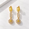 A pair of gold drop earrings with yellow rectangular stones, clear baguette side stones, and round yellow center stones, displayed on a white marble surface.