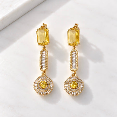 A pair of gold drop earrings with yellow rectangular stones, clear baguette side stones, and round yellow center stones, displayed on a white marble surface.