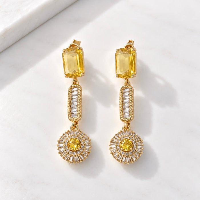A pair of gold drop earrings with yellow rectangular stones, clear baguette side stones, and round yellow center stones, displayed on a white marble surface.