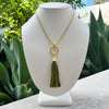 A gold chain necklace with a circular ring pendant and an olive green tassel, displayed on a white mannequin bust with a blurred garden background.