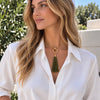 A woman wearing a gold-tone necklace with a green tassel pendant, styled with a white button-down shirt against an outdoor background of green foliage and a light stone wall.