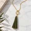 A gold chain necklace with a circular ring pendant and a large olive green tassel, styled on a white marble surface with a textured stone slab and plant leaves.