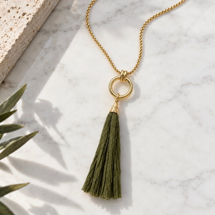 A gold chain necklace with a circular ring pendant and a large olive green tassel, styled on a white marble surface with a textured stone slab and plant leaves.