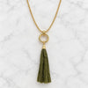 A gold chain necklace with a circular ring pendant and a hanging olive green fabric tassel, displayed on a white marble surface.