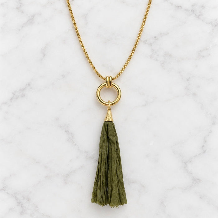 A gold chain necklace with a circular ring pendant and a hanging olive green fabric tassel, displayed on a white marble surface.