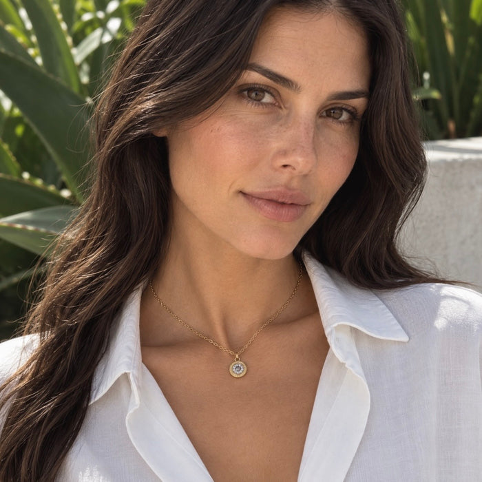 A woman with long dark brown hair wearing a delicate gold chain necklace with a circular pendant, styled with a white collared shirt, set against a soft-focus background of green foliage.