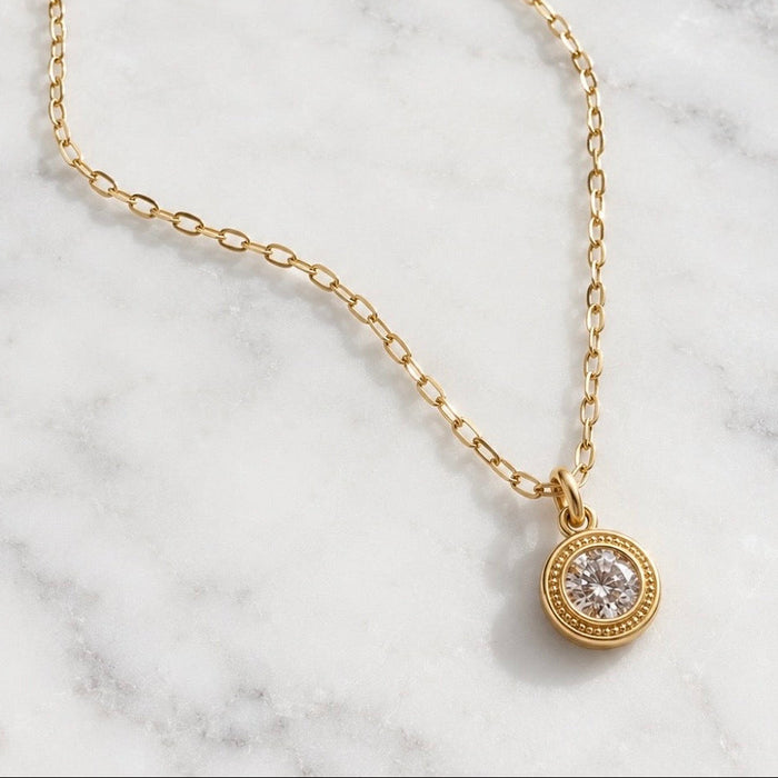 A delicate gold chain necklace with a small round pendant featuring a clear gemstone surrounded by a gold border, displayed on a white marble surface.