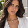 A close-up portrait of a woman with long dark brown hair wearing a delicate gold chain necklace with a small round pendant, paired with a white blouse. The background is a softly blurred outdoor setting.